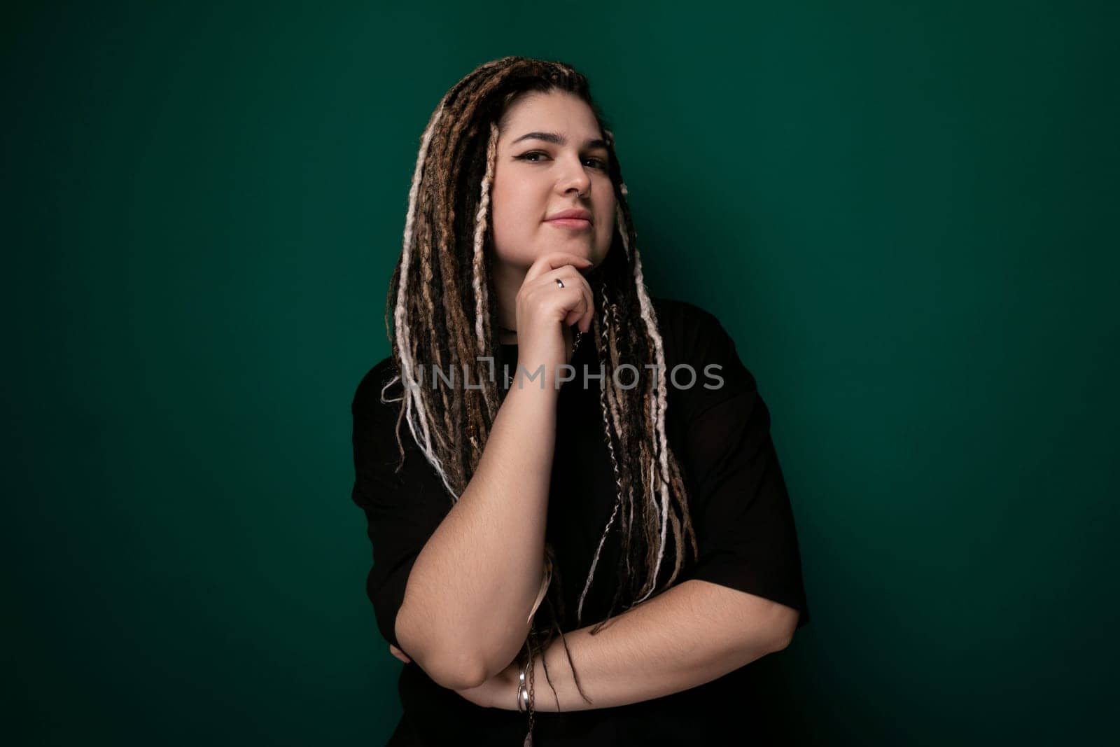 A woman with dreadlocks is standing in front of a vibrant green wall, looking directly at the camera. She appears confident and stylish in her outfit, with her hair cascading down her shoulders. The green wall provides a bold backdrop for the portrait.