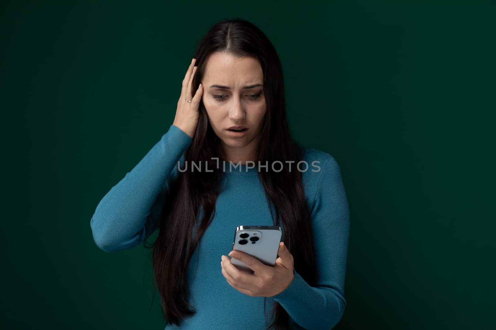 A woman wearing a blue shirt is seen holding a cell phone in her hand. She appears focused on the screen, perhaps texting or browsing. The background is neutral, allowing the woman and her phone to be the center of attention.