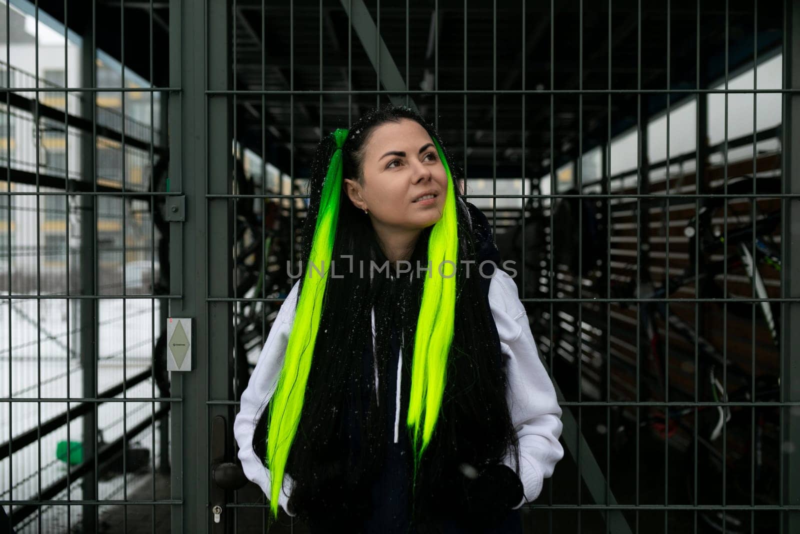 A woman with bright green hair stands confidently in front of a rustic wooden fence, her striking appearance contrasting with the natural surroundings. She gazes directly at the viewer, exuding a sense of strength and individuality.