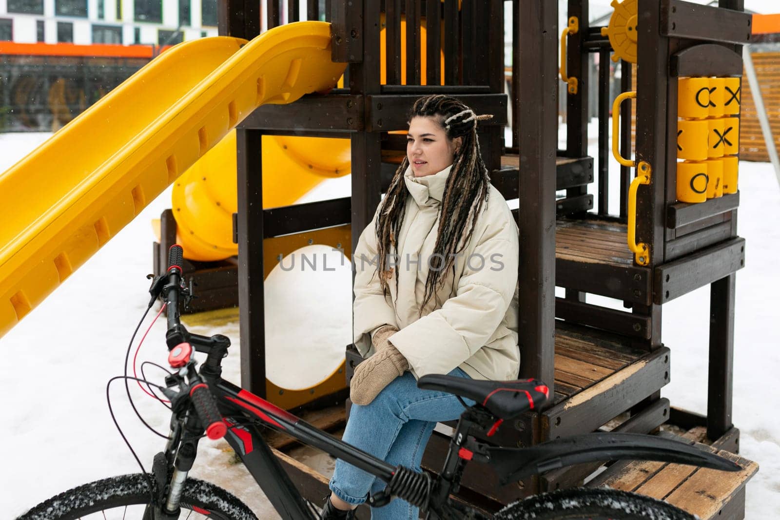 A woman is seated on a bench next to a parked bicycle in an urban setting. She appears to be taking a break or enjoying a moment of rest after cycling. The scene captures a simple, everyday moment of relaxation.