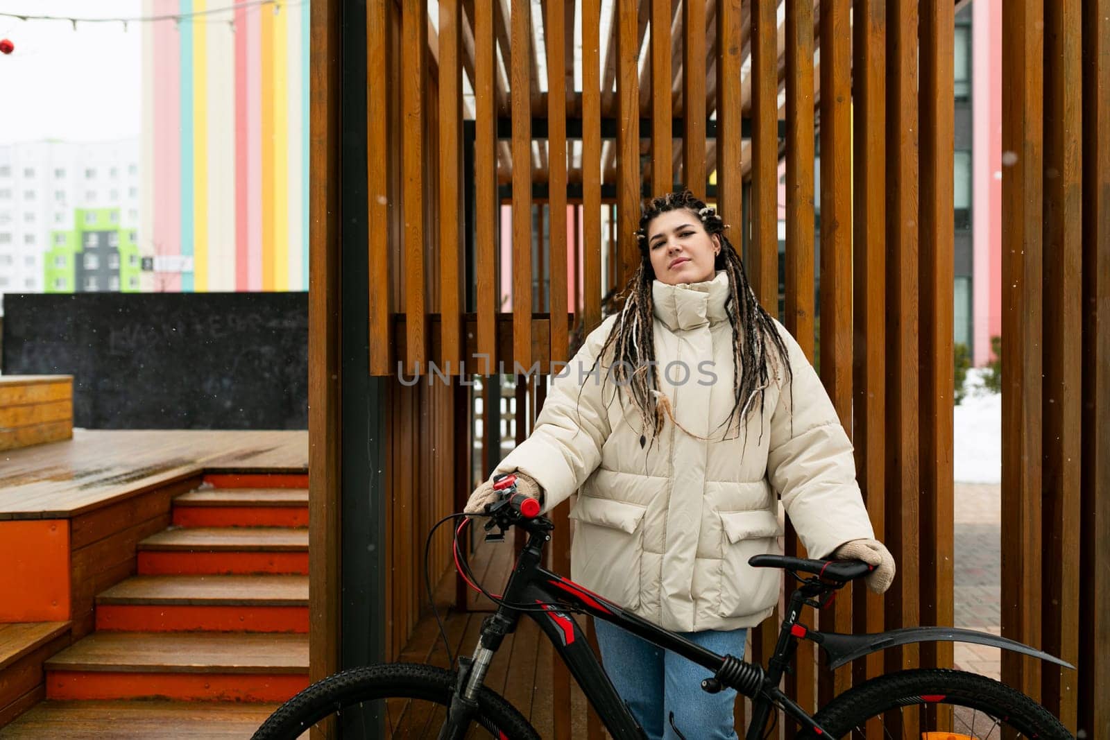 A woman with dreadlocks stands confidently next to her bicycle. She is wearing casual clothes and appears to be taking a break from cycling. The background shows an urban environment.