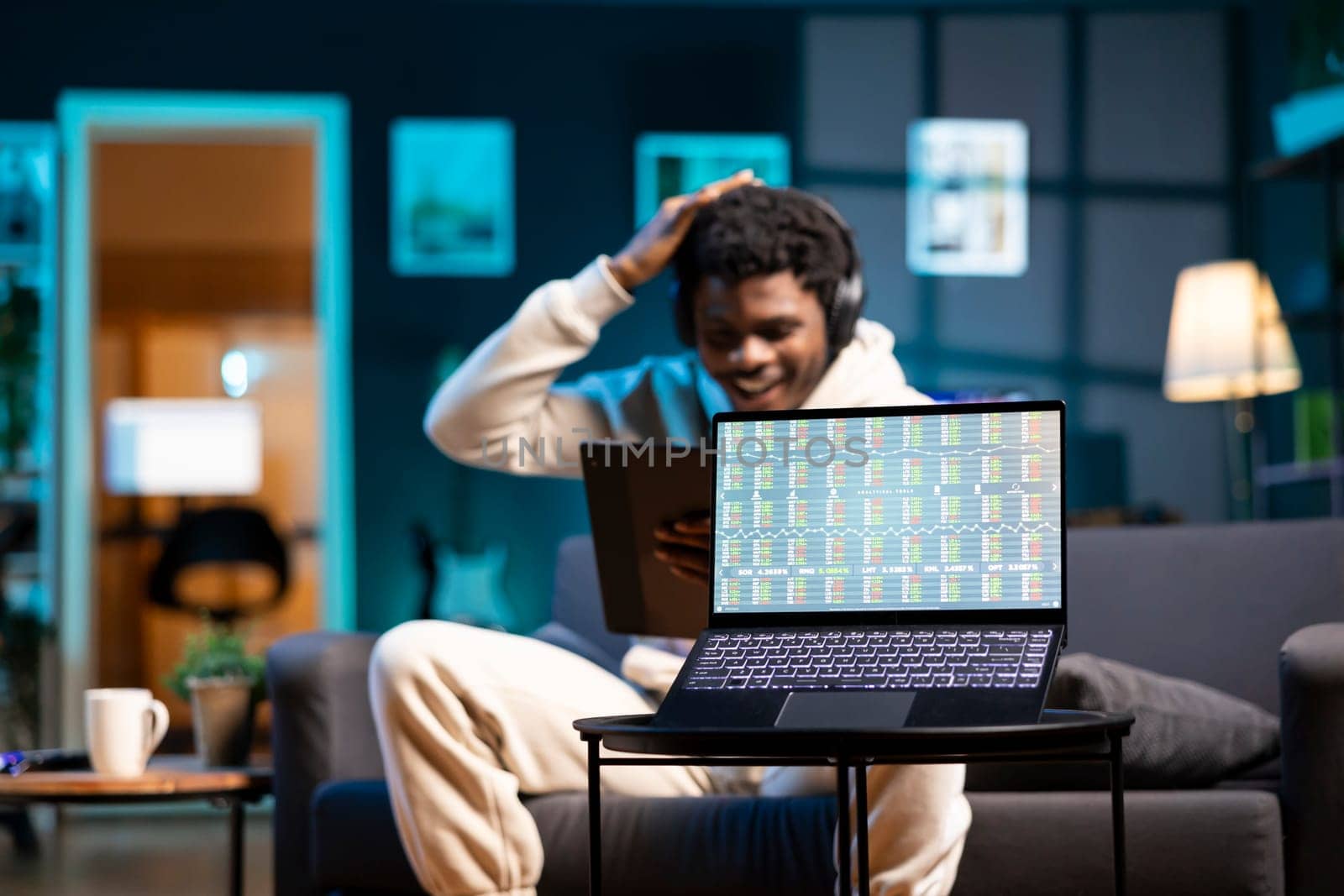 Smiling freelancer wearing headphones, analyzing investments on tablet, focus on stock market figures on laptop screen. Excited broker trader celebrating profit on trading charts