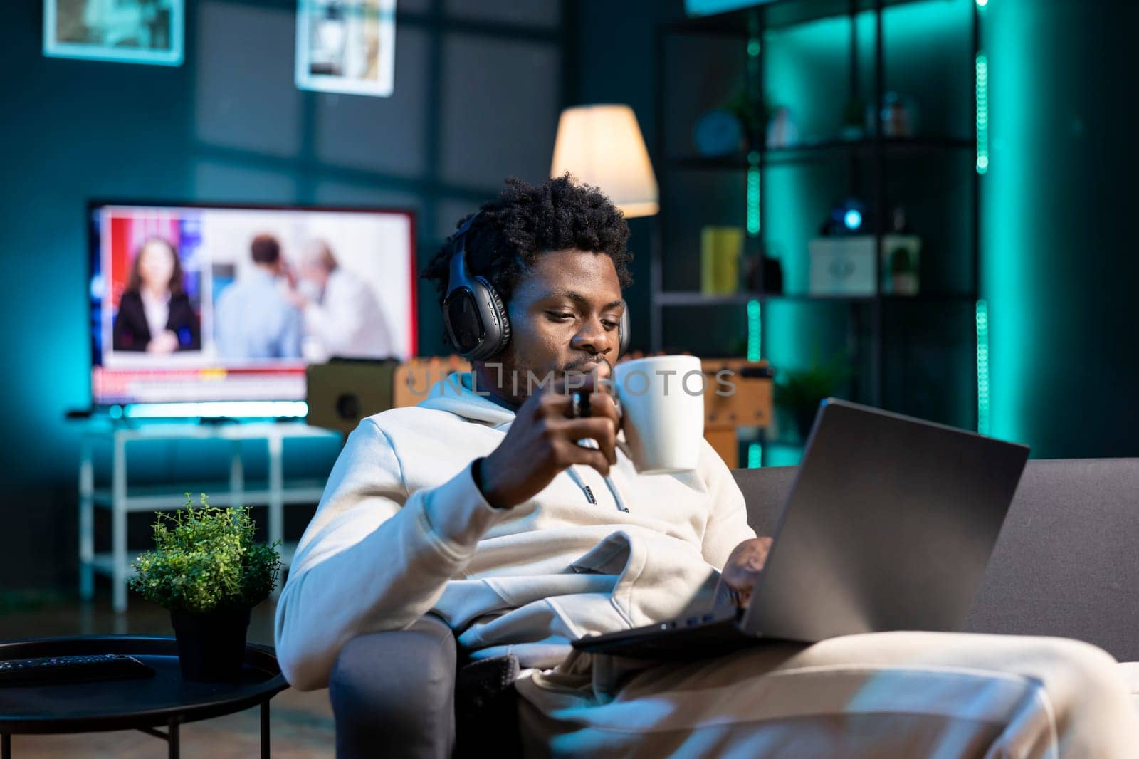 Happy man drinking coffee and writing emails at home office desk while listening music. Smiling remote worker doing b2b networking over internet, wearing headphones, enjoying hot beverage
