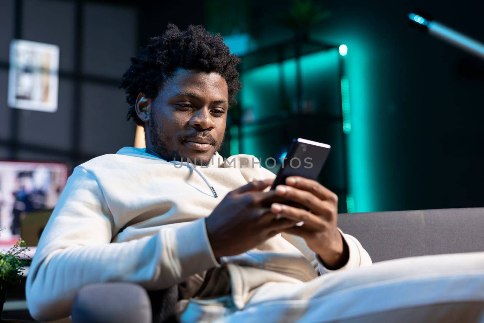 Smiling man impatiently texting friend, planning meeting hour together. Person sitting on cozy sofa in modern stylish apartment typing on cellphone, excited to meet up with mate