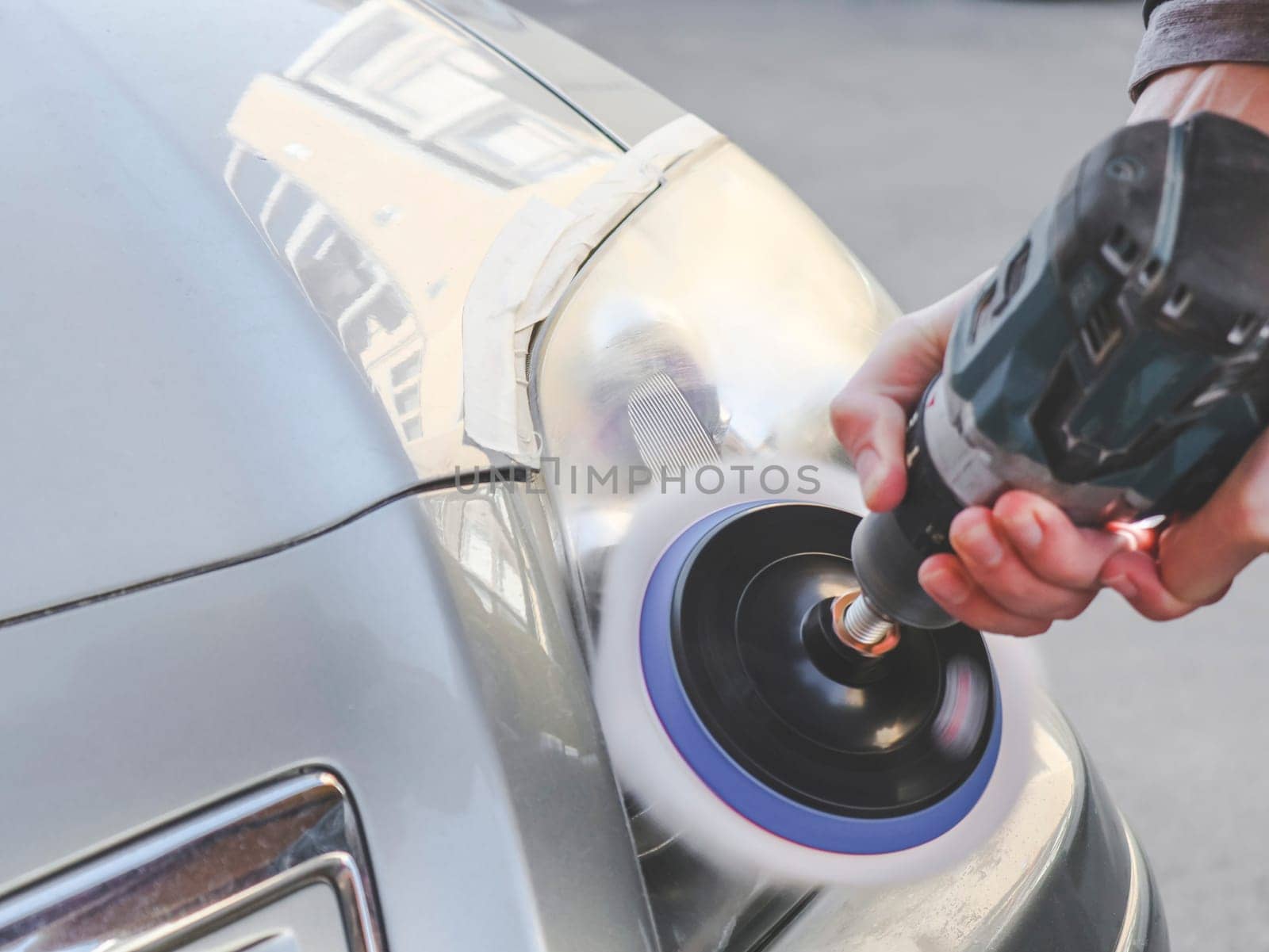 Hands of a young caucasian man in a yellow jacket polishing the headlights of a gray car with a washing soap with a drill and a sponge disk, close-up side view.Concept headlight polishing,car washing.