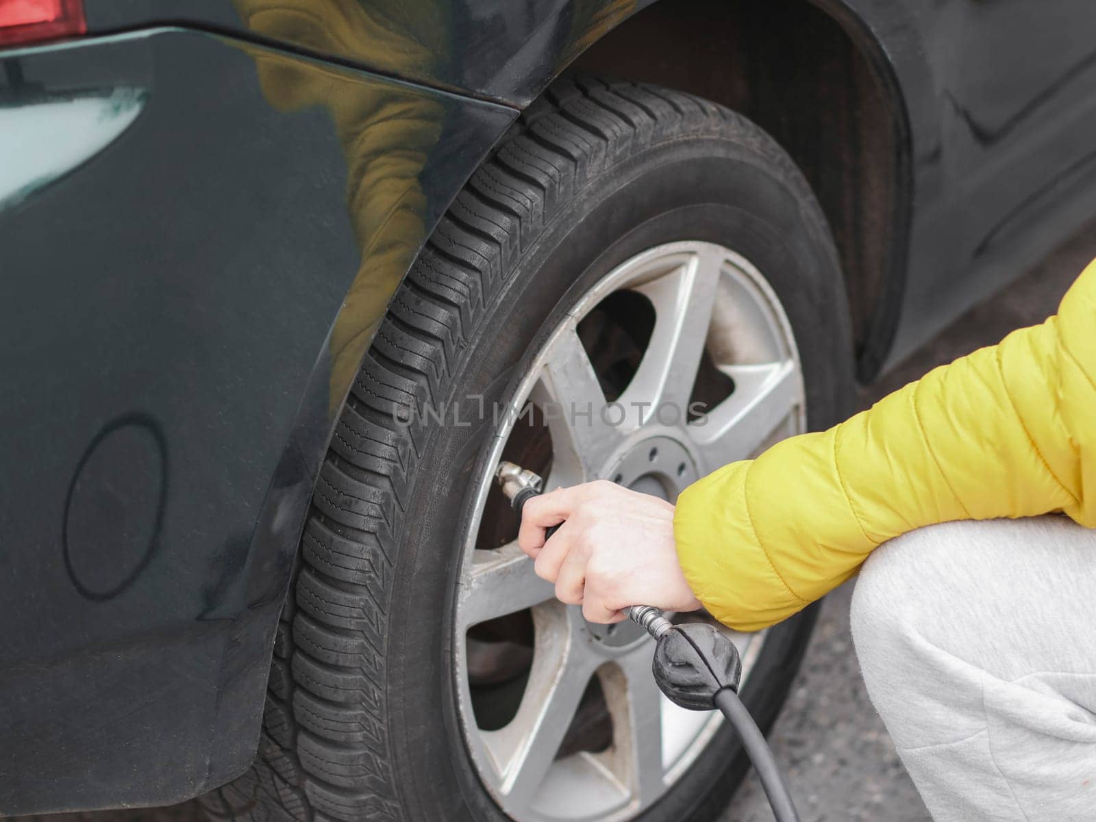 The hands of a young Caucasian man in a yellow jacket pump air with a compressor into the rear wheel of a car at a gas station, side view with selective focus and close-up. Gas station concept.