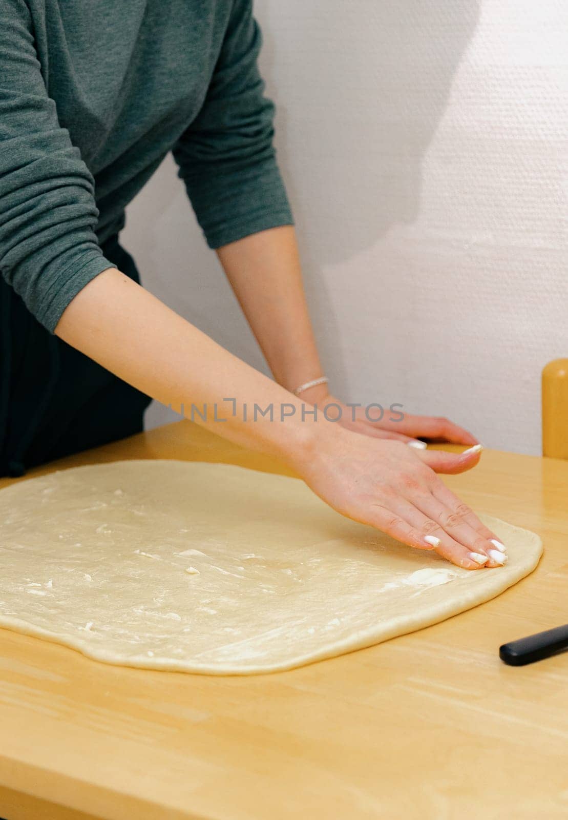 One unrecognizable young Caucasian girl spreads butter with her fingers onto the yeast dough on a wooden table, standing in the kitchen, close-up side view. Step-by-step instructions for baking synabons. Step 6.