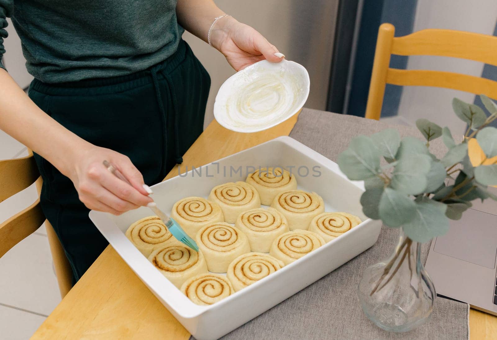 One unrecognizable young Caucasian girl butters round uncooked buns with brown sugar and cinnamon in a white baking dish on a wooden table while standing in the kitchen, close-up side view. Step-by-step instructions for baking synabons. Step 12