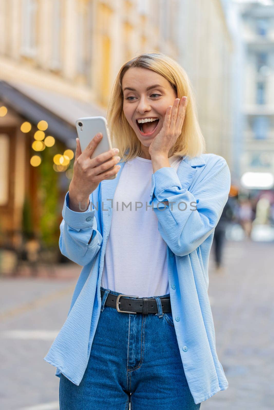 Caucasian young woman using mobile smartphone celebrating win good message news, lottery jackpot victory, giveaway online outdoors. Happy adult lady tourist standing in city street. Town lifestyles