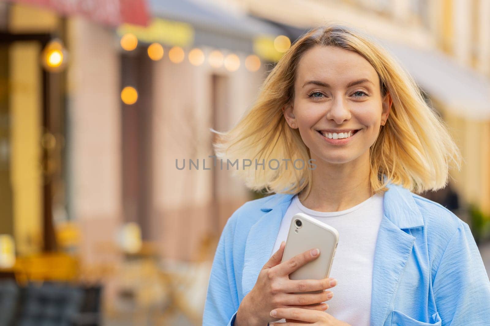 Smiling young woman using smartphone typing text messages browsing internet, finishing work, looking at camera outdoors. Lovely lady on urban city street. Camera zooming in and person becomes in focus