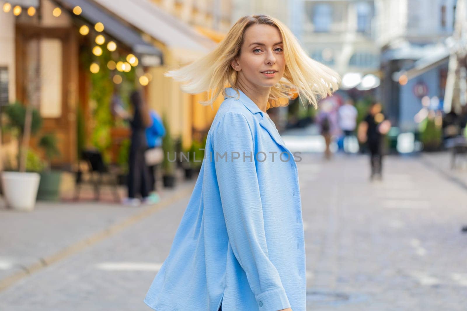 Rear view of young happy blonde woman tourist walking through European street outdoors turns and looking at camera. Back view of lovely pretty girl lady traveler walk in urban city. Town lifestyles