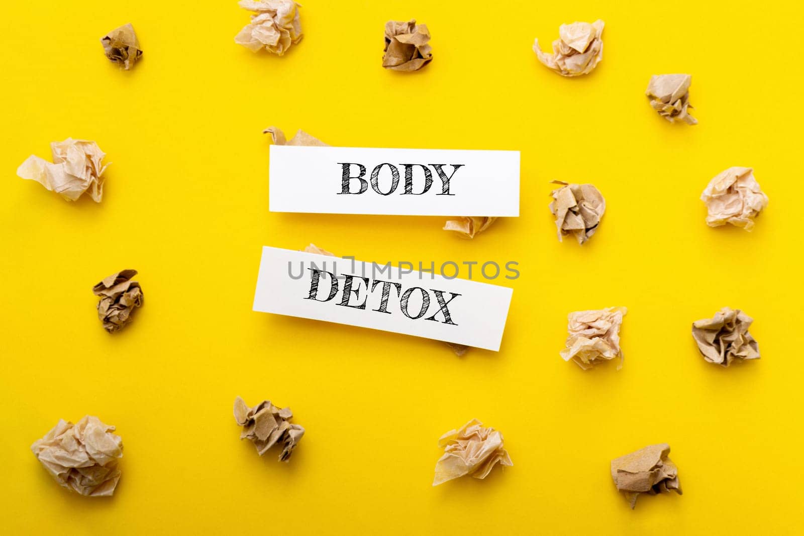 Two pieces of paper with the words body detox written on them. The paper is placed on top of a yellow background, which is covered in crumpled paper. Scene is somewhat chaotic