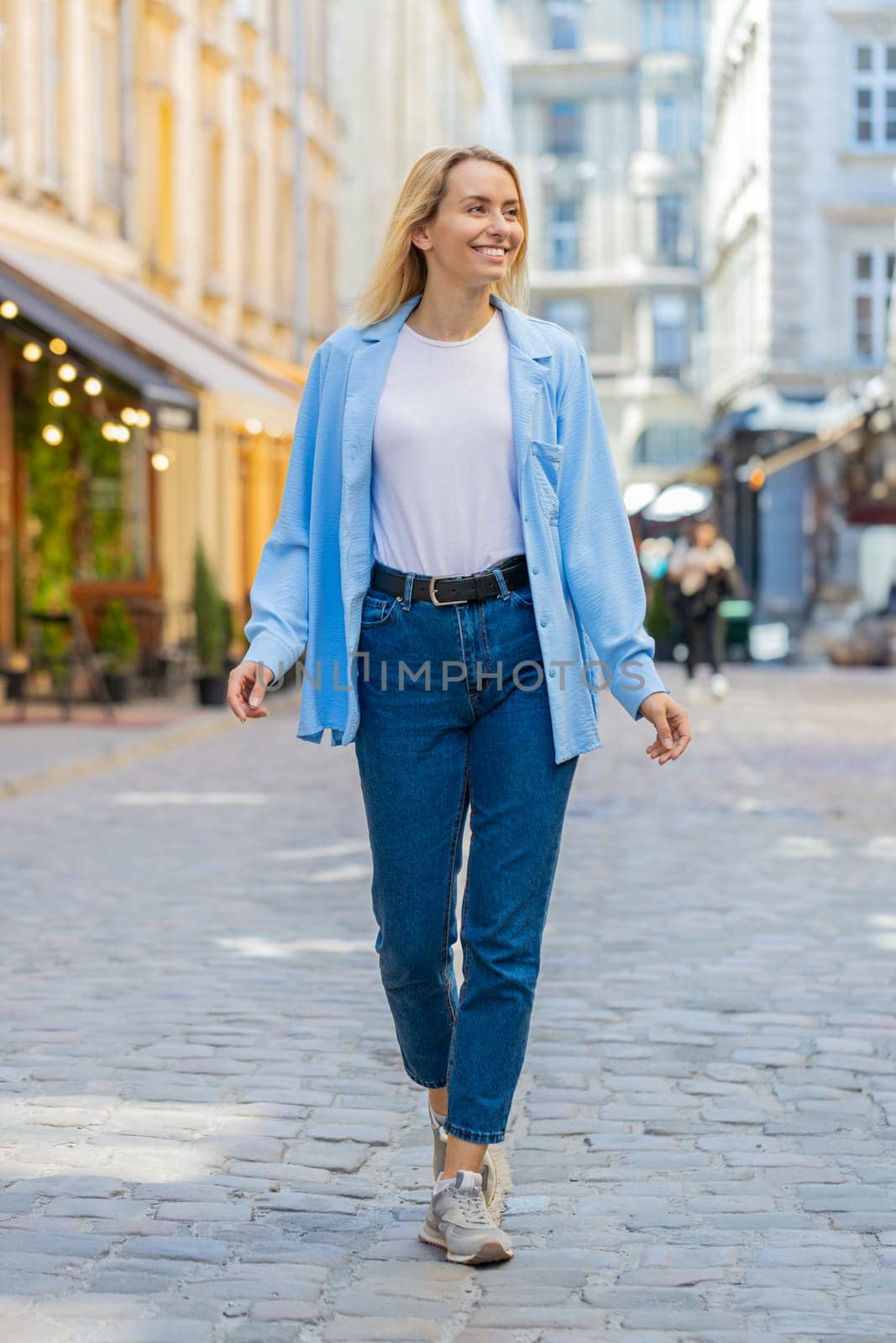 Portrait of young blonde tourist woman walking on urban city street outdoors. Smiling happy lady girl traveler having positive good mood enjoying in summer daytime. Town lifestyles, vacation holidays