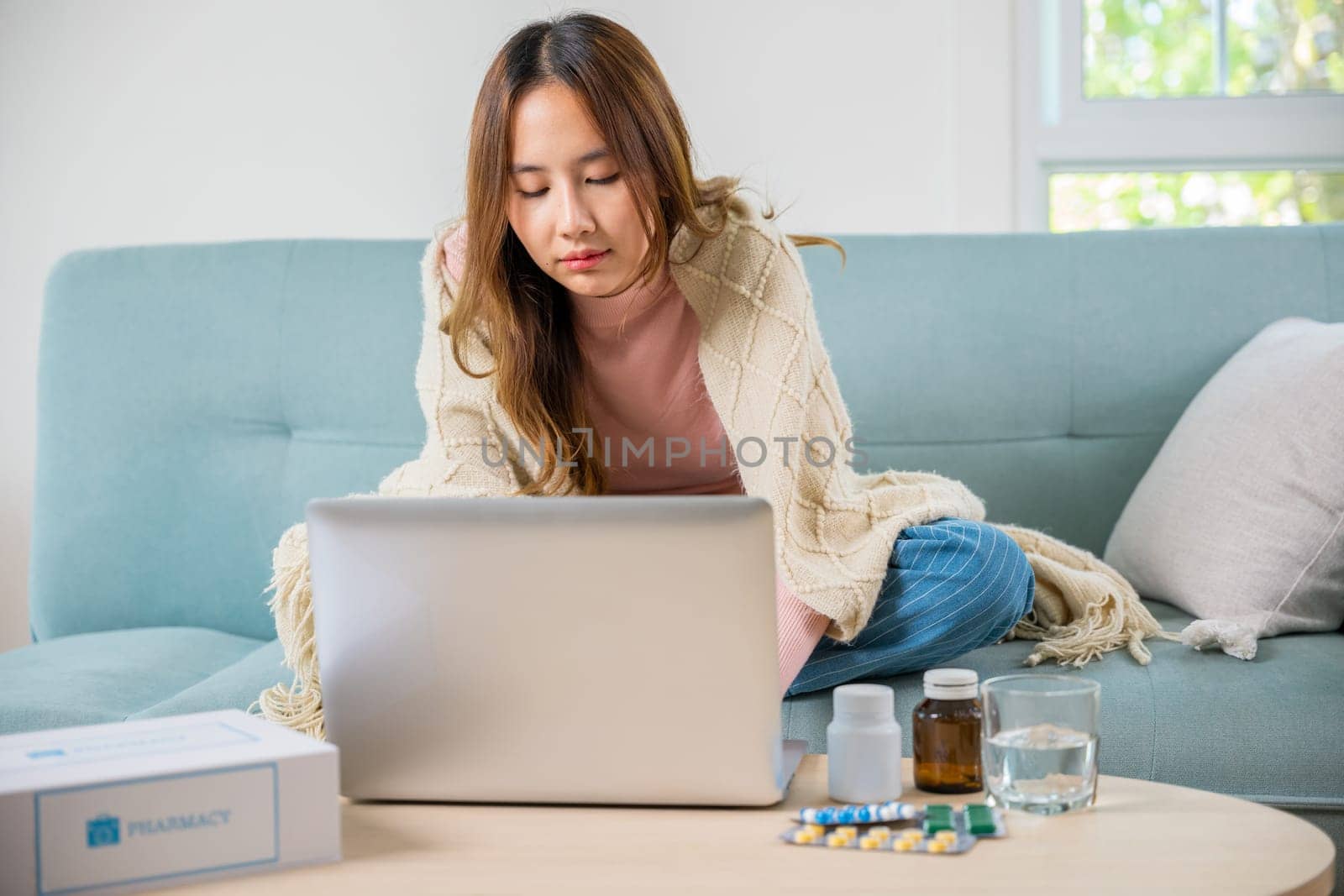 Business female sick she taking pills for flu while work with laptop keyboard, Asian sick woman covered blanket suffering from covid-19 virus but still working at home with laptop computer