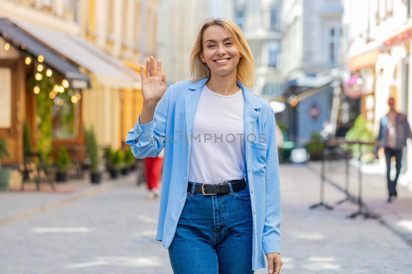 Cheerful young woman tourist smiling friendly at camera, waving hands gesturing invitation hello, hi, greeting, goodbye, welcoming with hospitable expression outdoors. Lady on urban city town street