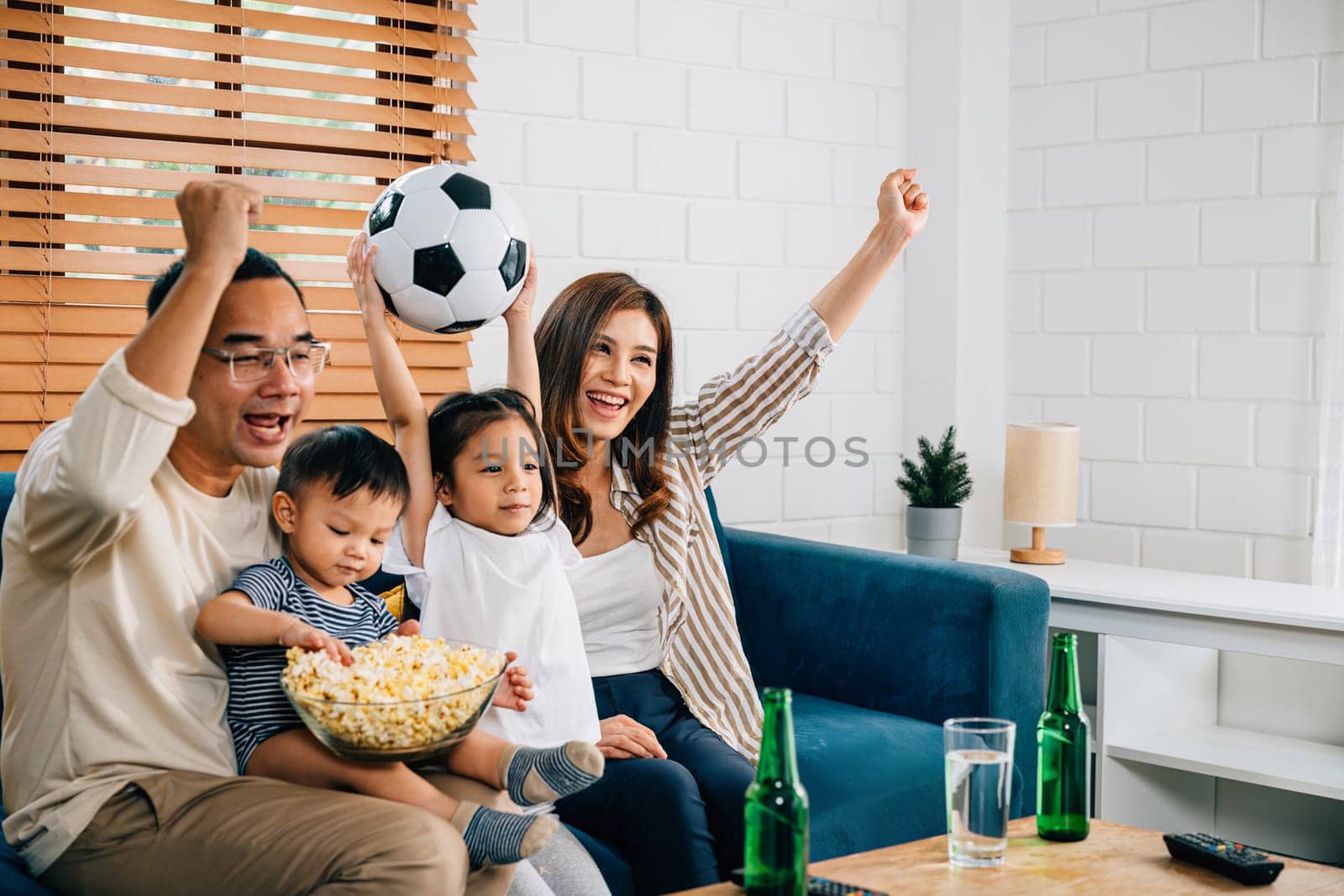 A jubilant family watches a football match with popcorn and a ball, raising arms and celebrating. Their togetherness and cheers reflect the joy of winning and their shared love for the sport.