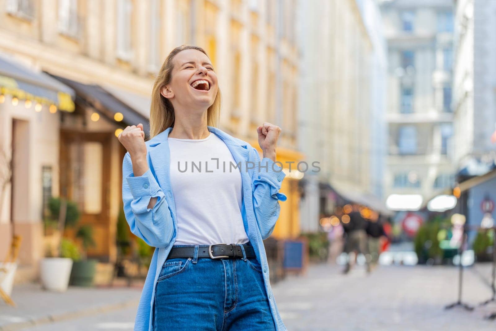 Excited happy woman traveler shouting, celebrating success, winning, goal achievement, good news, lottery jackpot luck, victory outdoors. Young lady walking on urban city sunshine street. Lifestyles