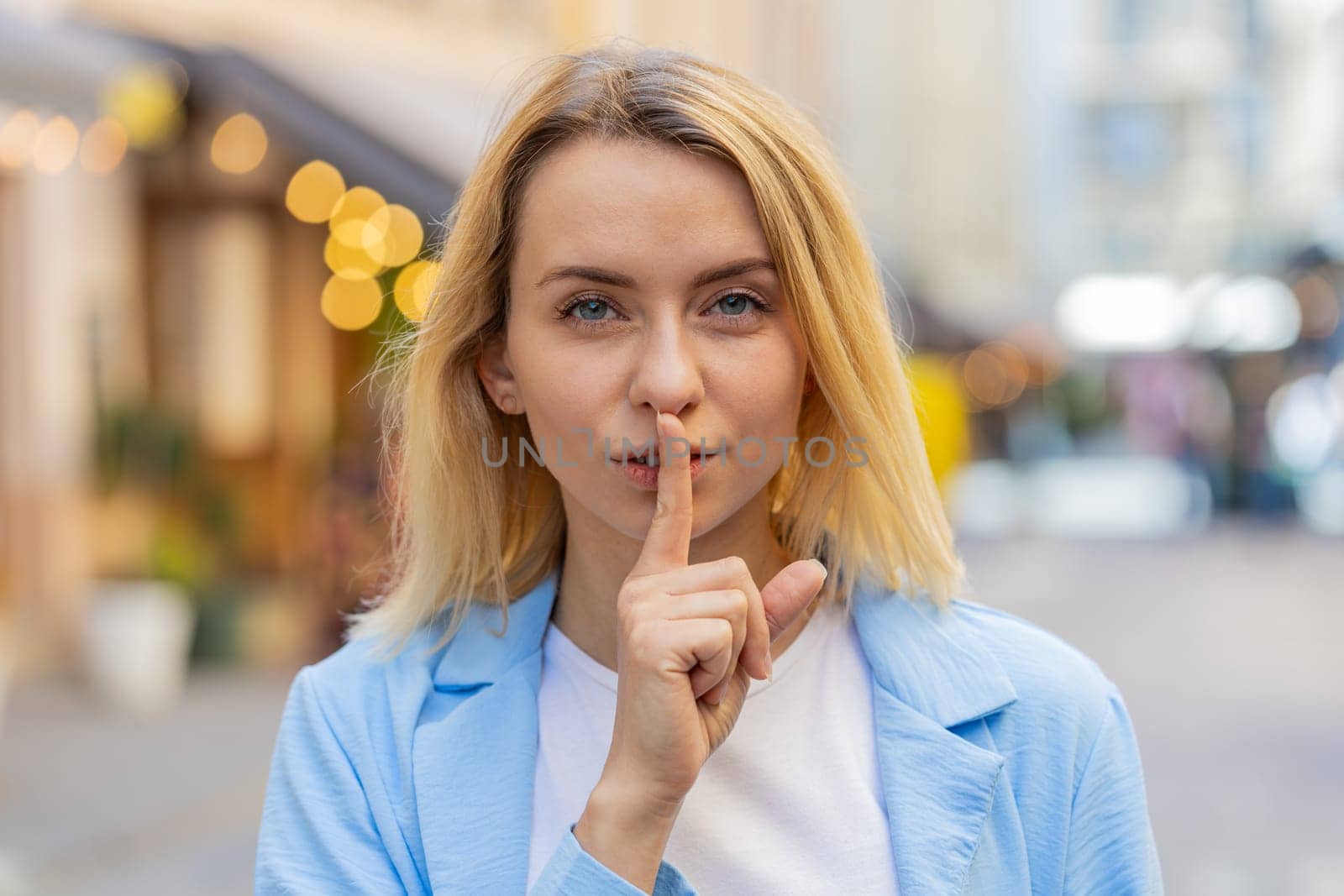 Shh be quiet please. Blonde young woman presses index finger to lips makes silence hush gesture sign do not tells gossip secret outdoors. Young lady female stand on urban city street. Town lifestyles