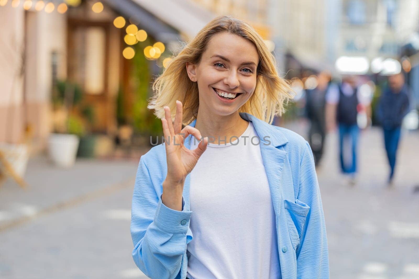 Okay. Happy cheerful young Caucasian woman looking approvingly at camera showing OK gesture positive like sign, approve something good celebrate win. Joyful lady standing in urban city street outdoors