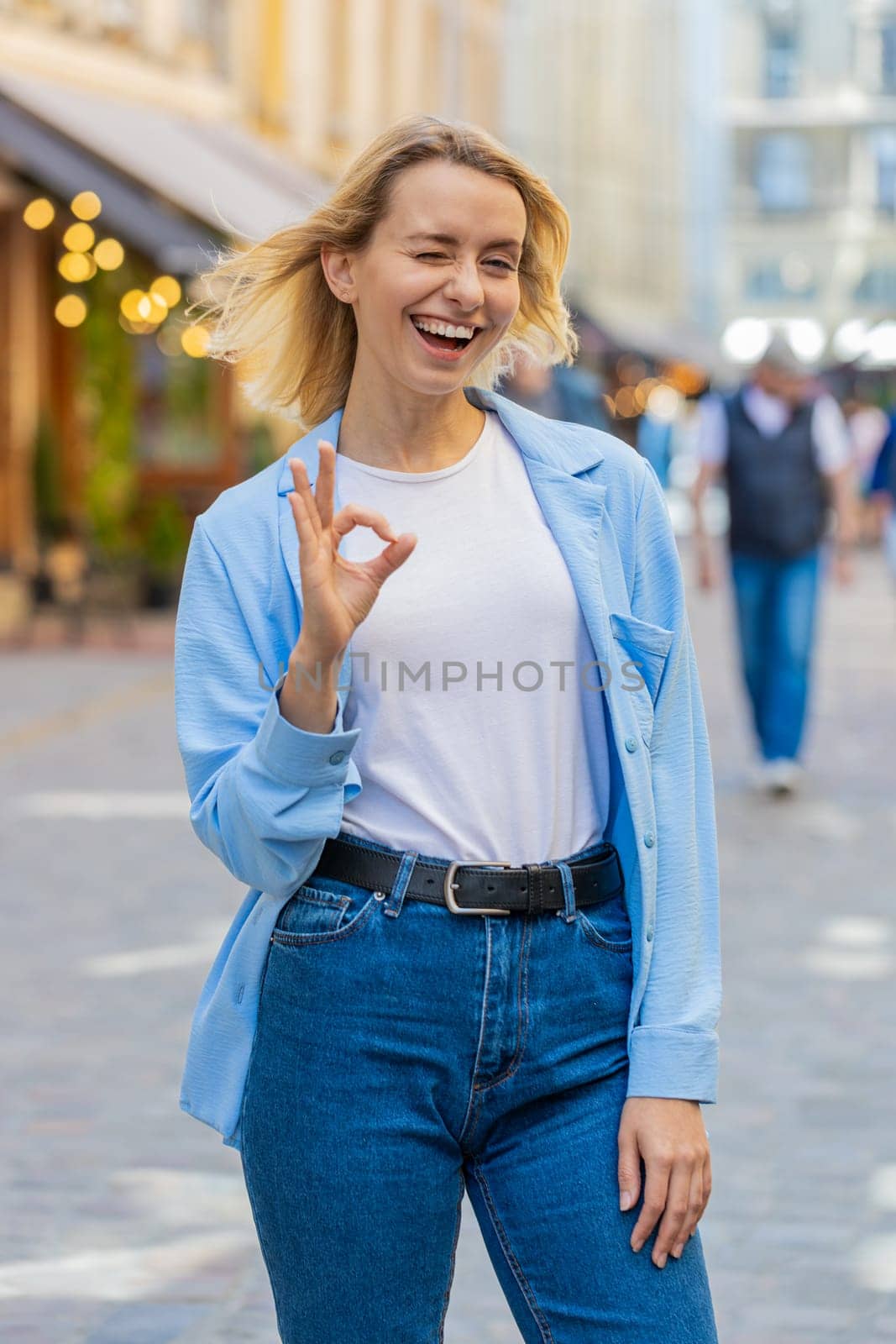 Okay. Happy cheerful young Caucasian woman looking approvingly at camera showing OK gesture positive like sign, approve something good celebrate win. Joyful lady standing in urban city street outdoors