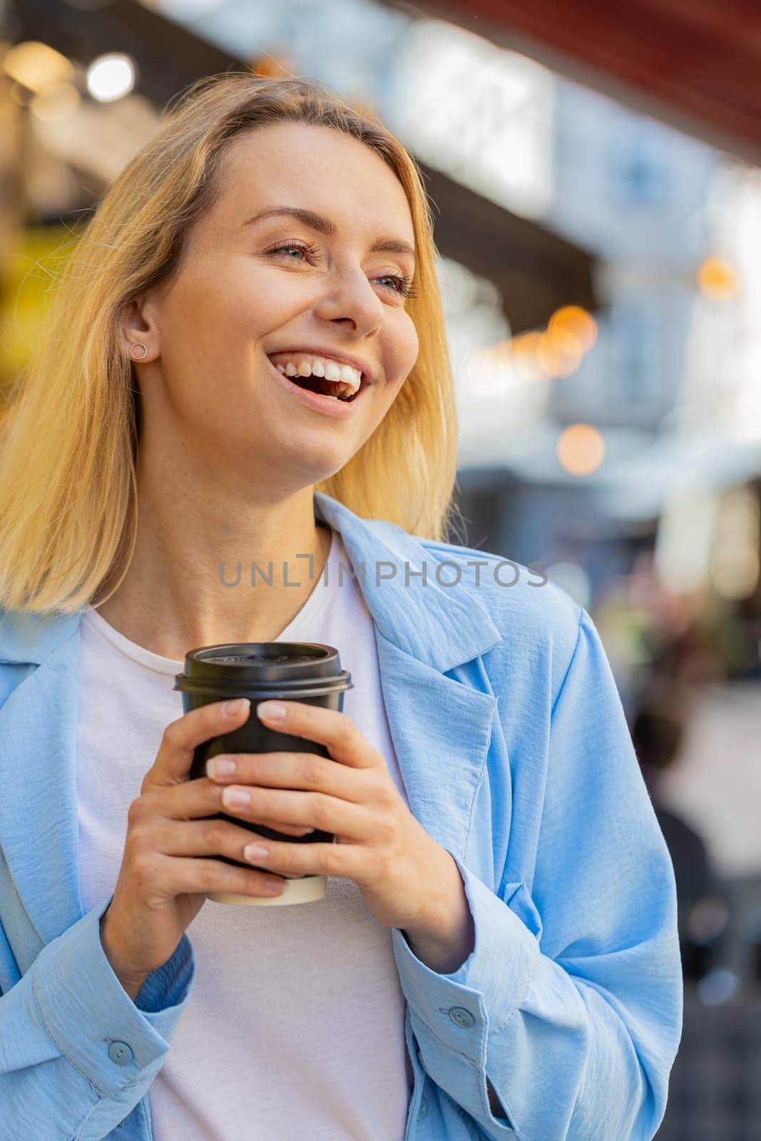 Happy Caucasian adult woman enjoying morning coffee hot drink and smiling. Relaxing, taking a break. Young lady standing on urban city center street, drinking coffee to go. Town lifestyles outside.