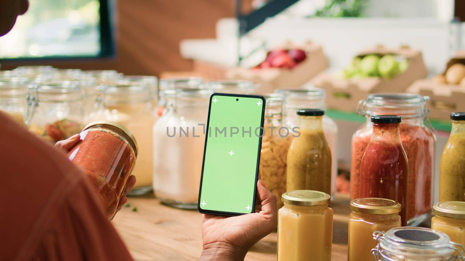 African american client uses greenscreen while she is looking at organic eco bulk products stored in reusable jars. Woman customer checks smartphone with isolated chromakey mockup display.