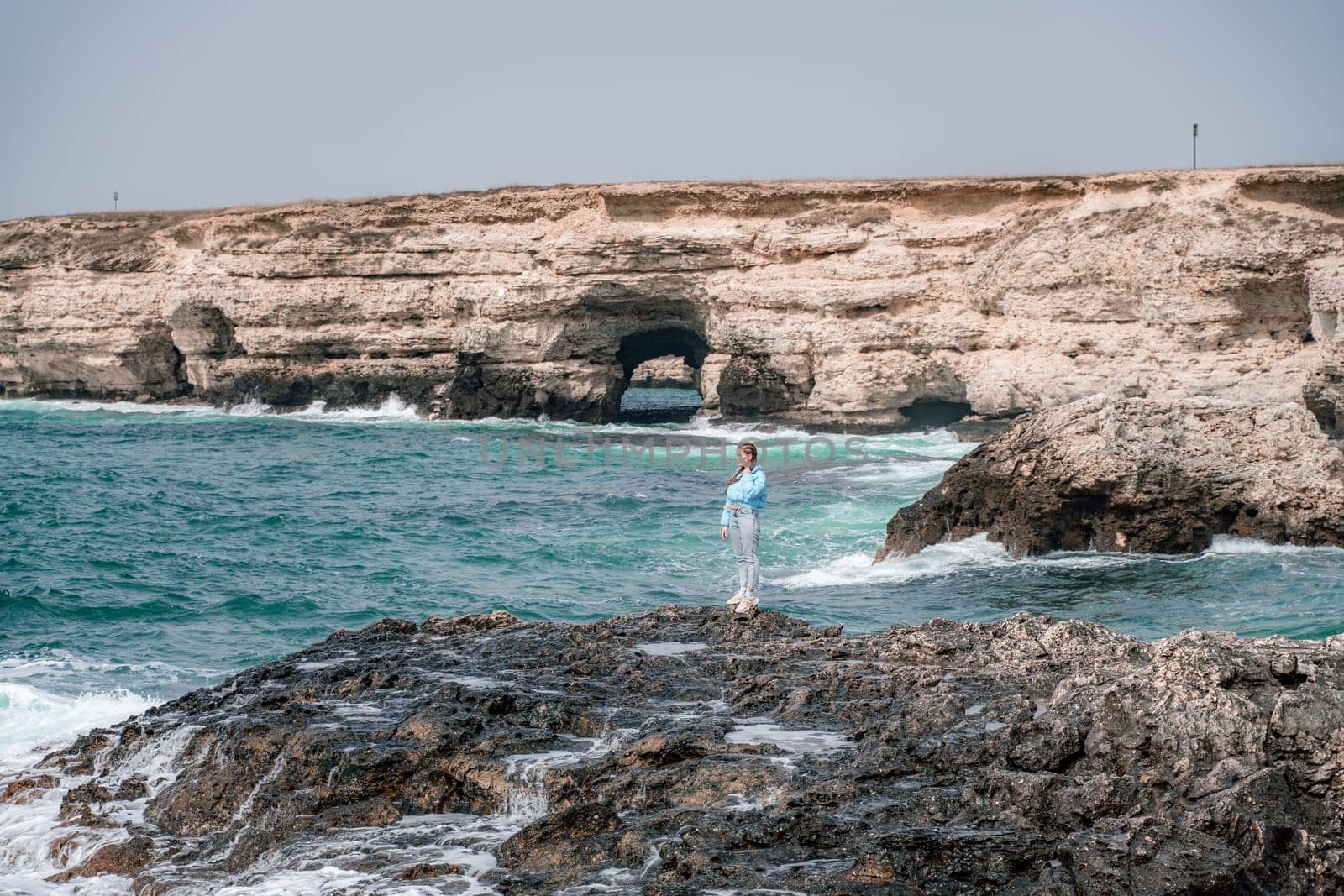 A woman in a blue jacket stands on a rock above a cliff above the sea and looks at the raging ocean. Girl traveler rests, thinks, dreams, enjoys nature. Peace and calm landscape, windy weather