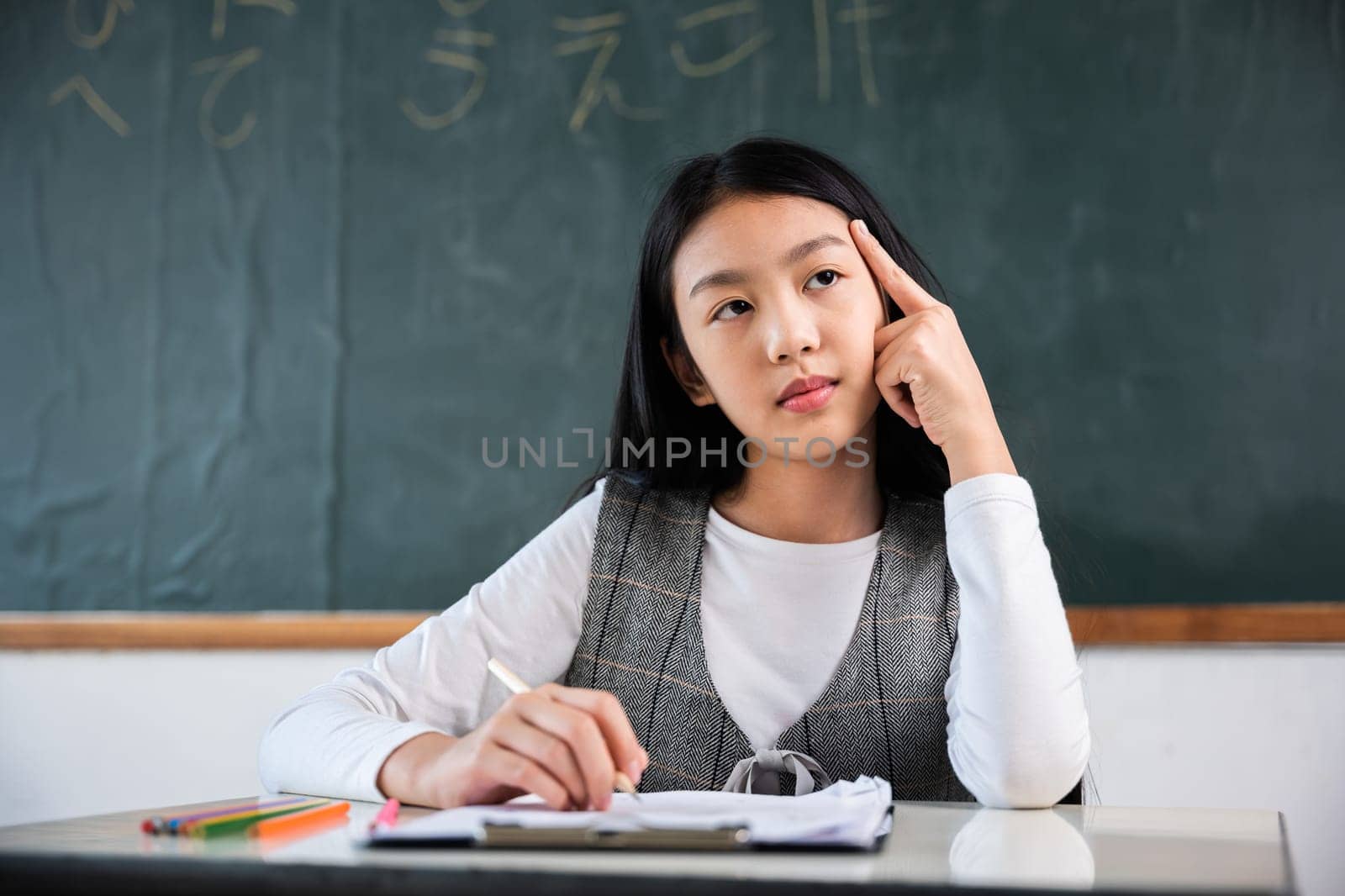 A girl is sitting at a desk in a classroom and is writing in a notebook by Sorapop