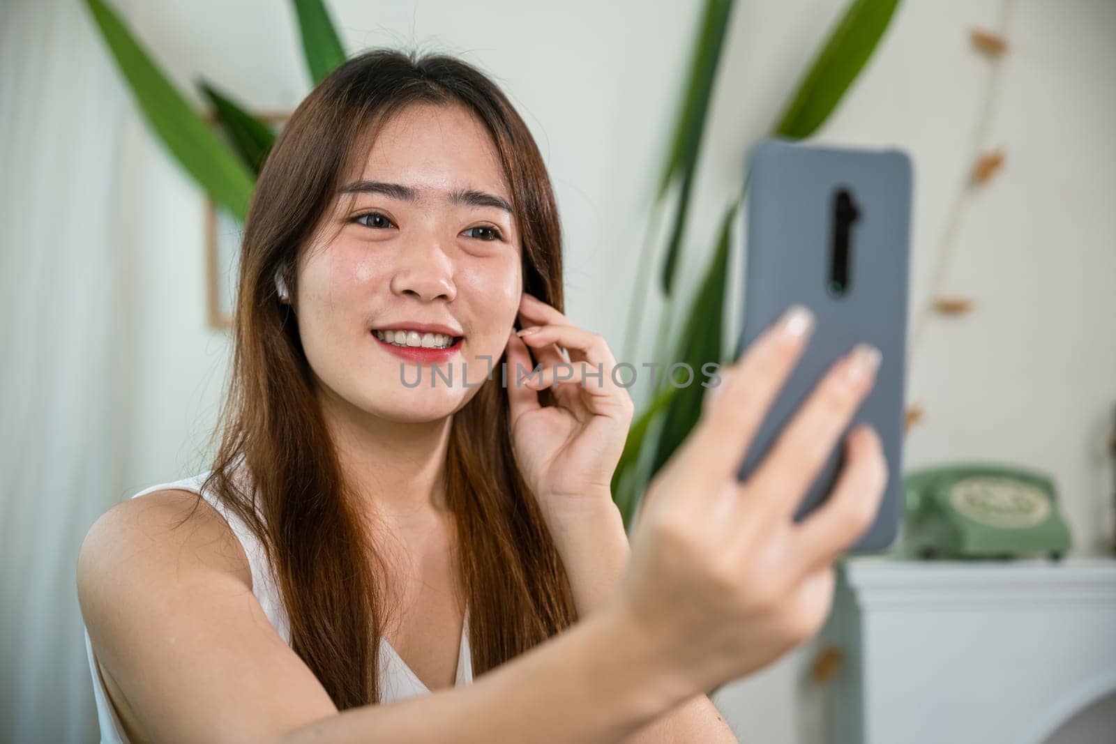 Happy relaxed female touching earbuds listen song on mobile phone in bright living room at home, Lifestyle Asian young woman wearing wireless earphones listening to favorite song on her smartphone