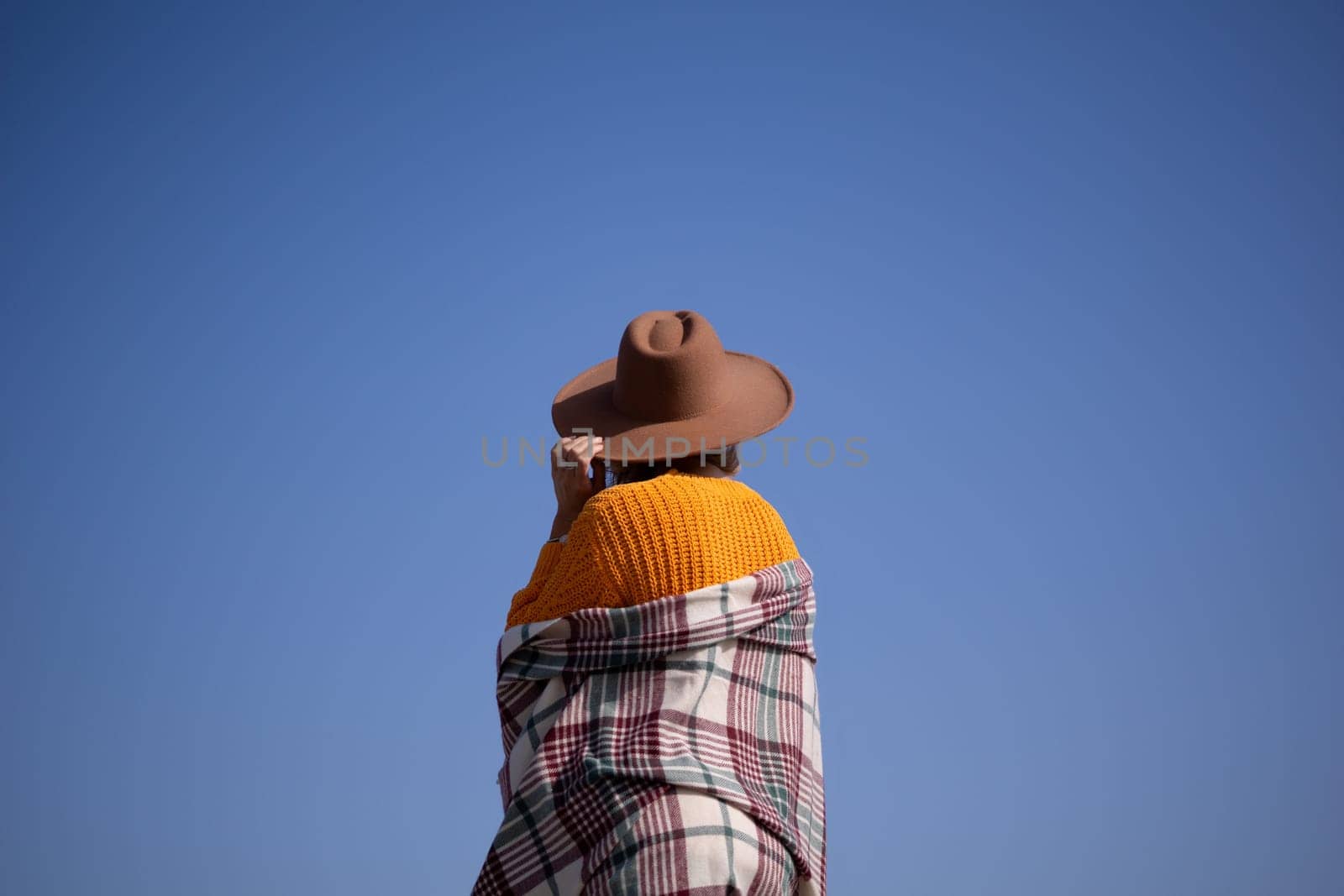 A woman wearing a brown hat and a plaid blanket stands in front of a blue sky. Concept of warmth and comfort, as the woman is wrapped in a cozy blanket
