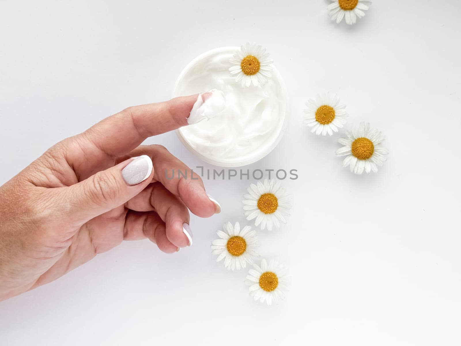 Close up of adult middle age woman hand with cream on finger, chamomile flowers, and hand cream container on white background. Skincare and natural beauty concept. Healthcare, wellness, and organic.