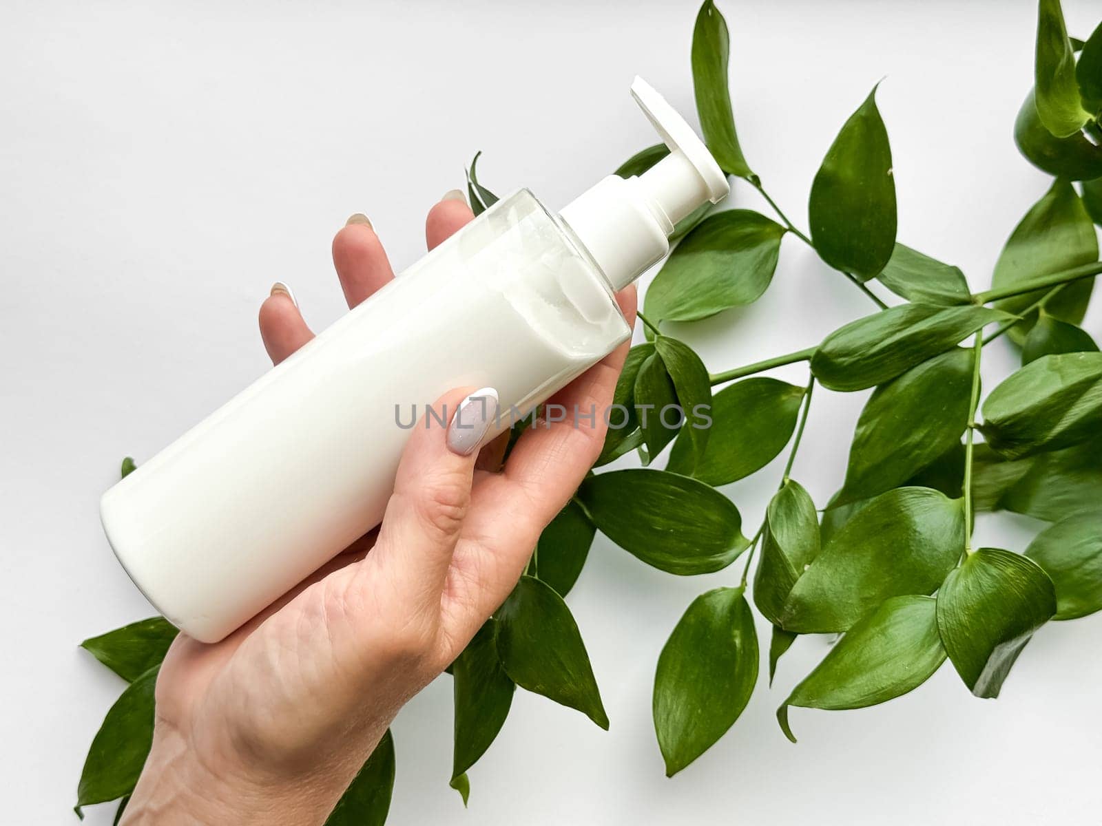 Close up of middle age woman hand holding white lotion bottle with green leaves on white background. Skincare and natural beauty concept. Healthcare, wellness, and organic product poster. High quality