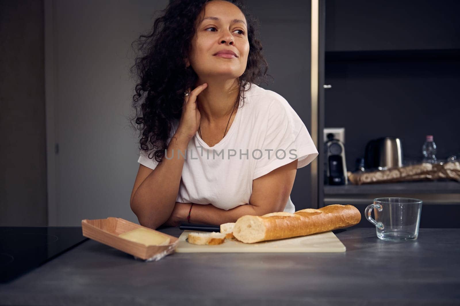 Authentic beautiful curly haired brunette woman in white t-shirt, dreamily looking aside, standing at kitchen countertop with a loaf of fresh bread and slice of cheese for breakfast. People. Lifestyle