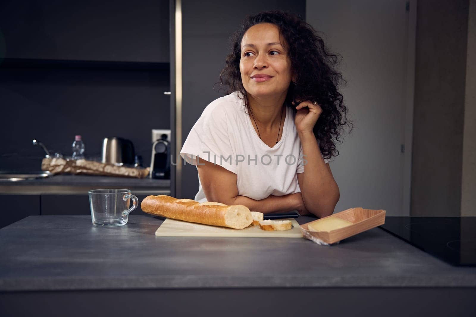 Curly haired multi ethnic beautiful brunette woman in white t-shirt, dreamily looking aside, standing at kitchen countertop with a loaf of bread and slices of cheese for breakfast. People. Lifestyle