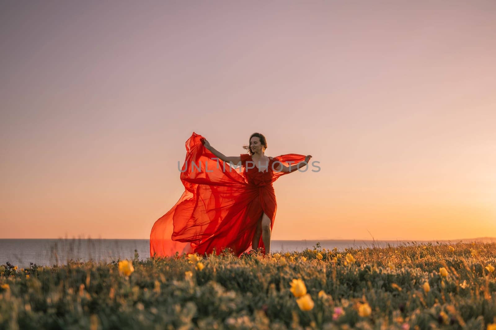 woman red dress is standing on a grassy hill overlooking the ocean. The sky is a beautiful mix of orange and pink hues, creating a serene and romantic atmosphere