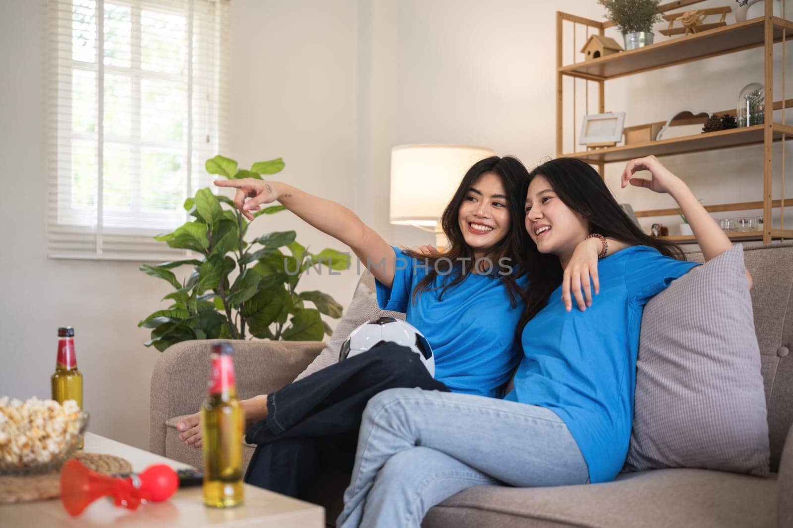 A lesbian couple cheers football and celebrate together for their favorite Euro football team. A young female couple cheers football on TV together in the living room on match day. by wichayada