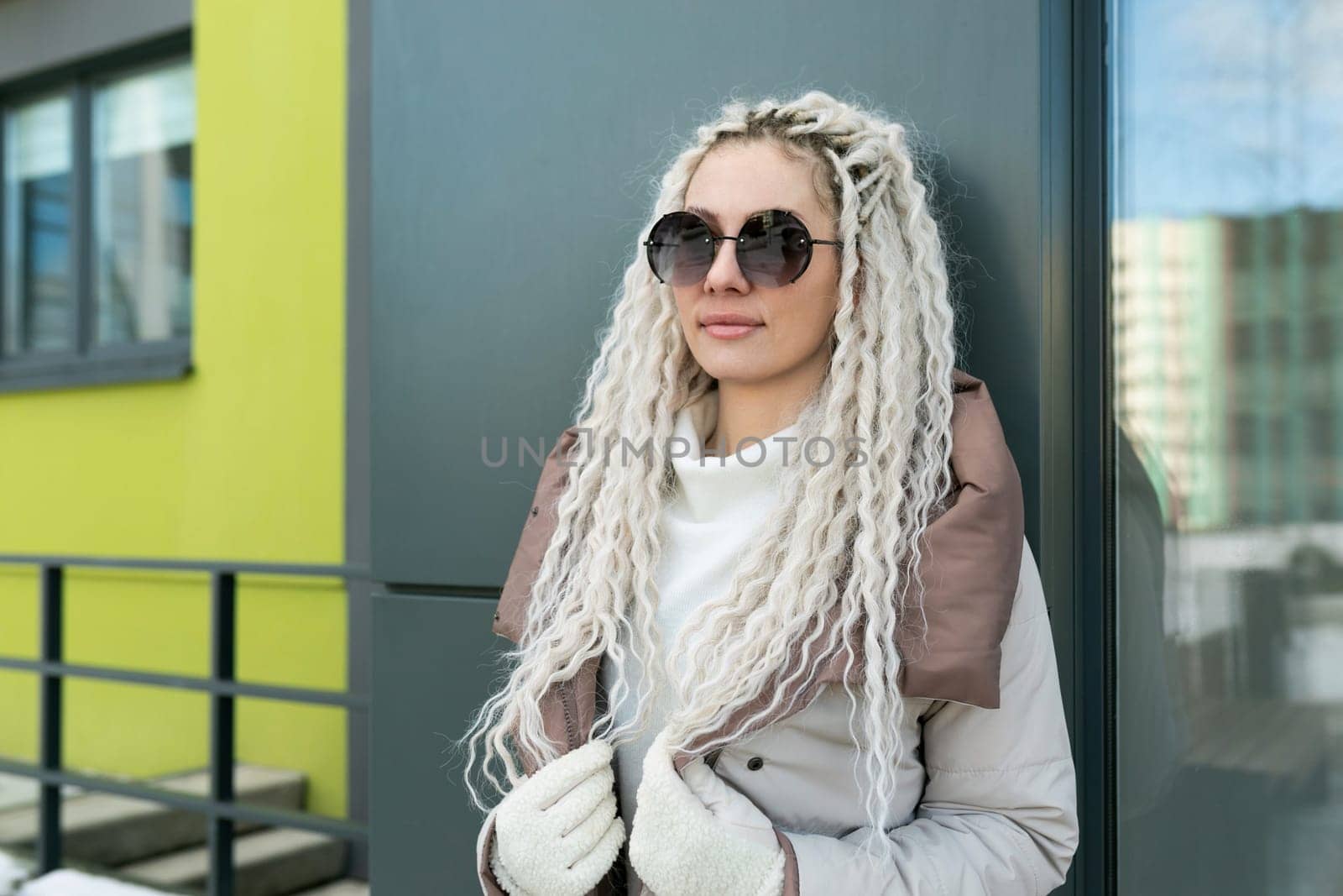 A woman with white dreadlocks leaning against a wall in an urban setting. She is dressed casually, with a contemplative expression on her face.