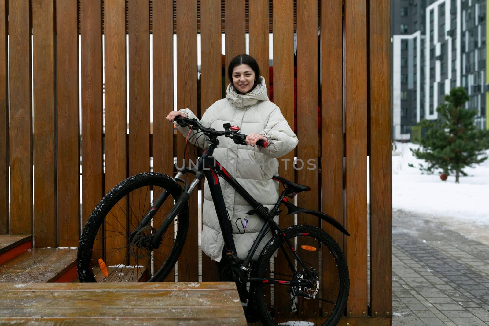 A woman is standing next to a bike in front of a wooden fence. She appears to be adjusting something on the bike. The setting is outdoors in a residential area.