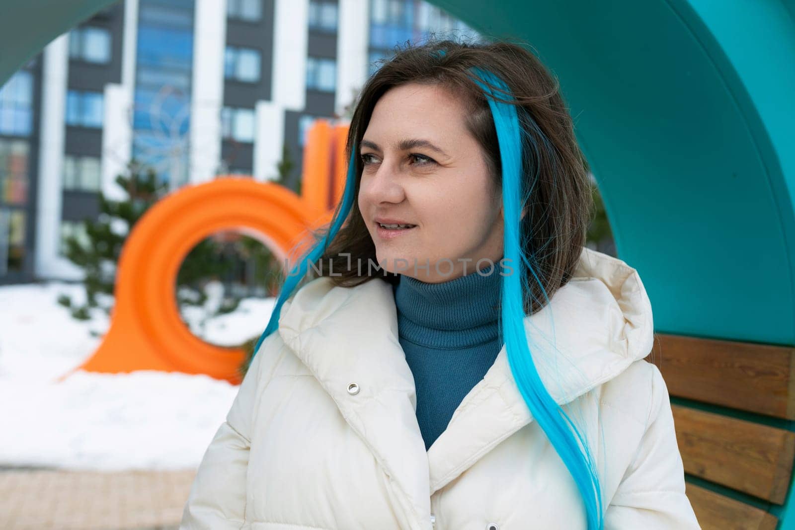 A woman with vibrant blue hair sits on a wooden bench in a public park. She is dressed in casual clothing, with headphones in her ears, looking off into the distance.