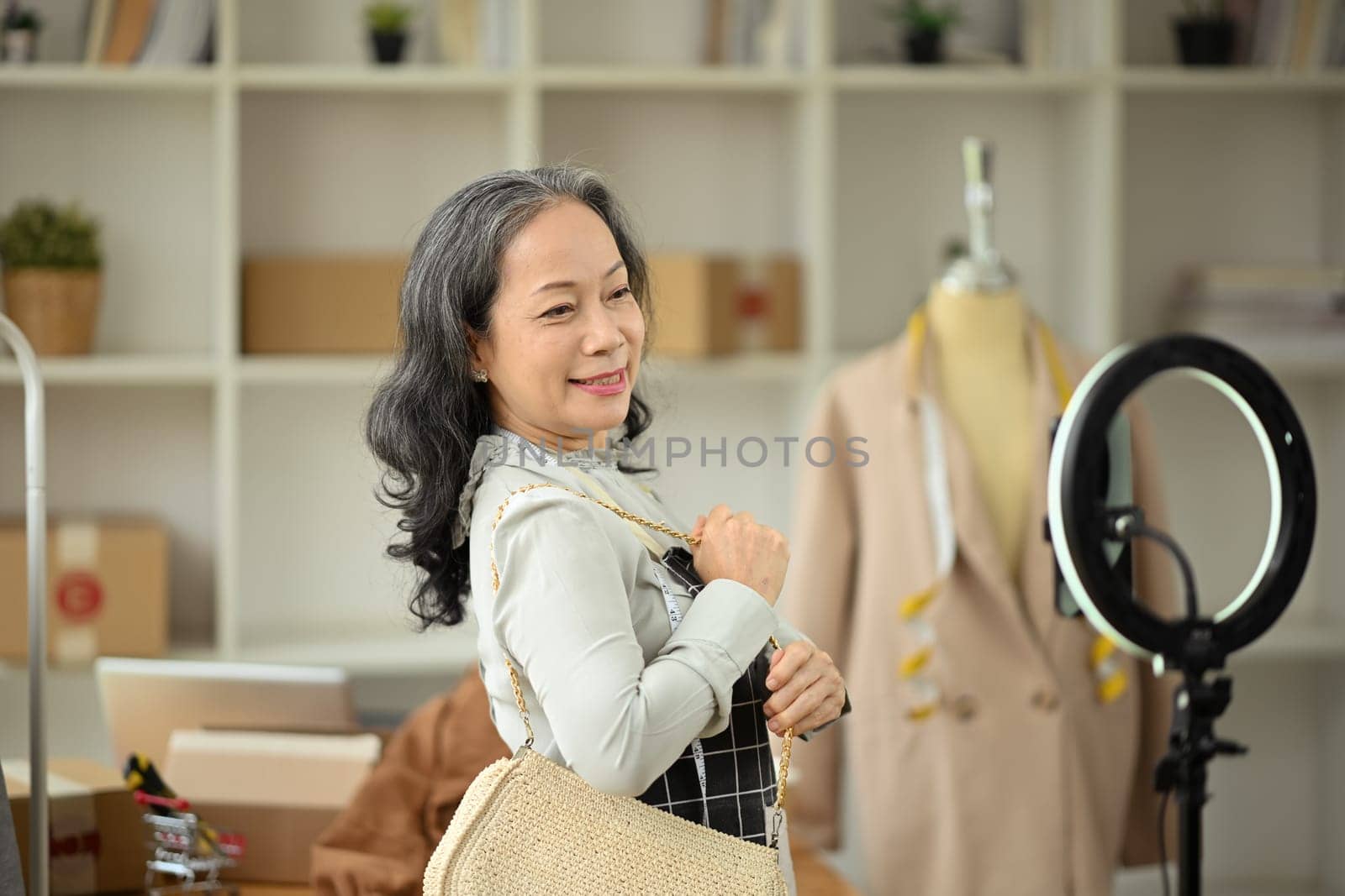 Cheerful senior female business owner showing handbag in front of smart phone, selling product on live stream.