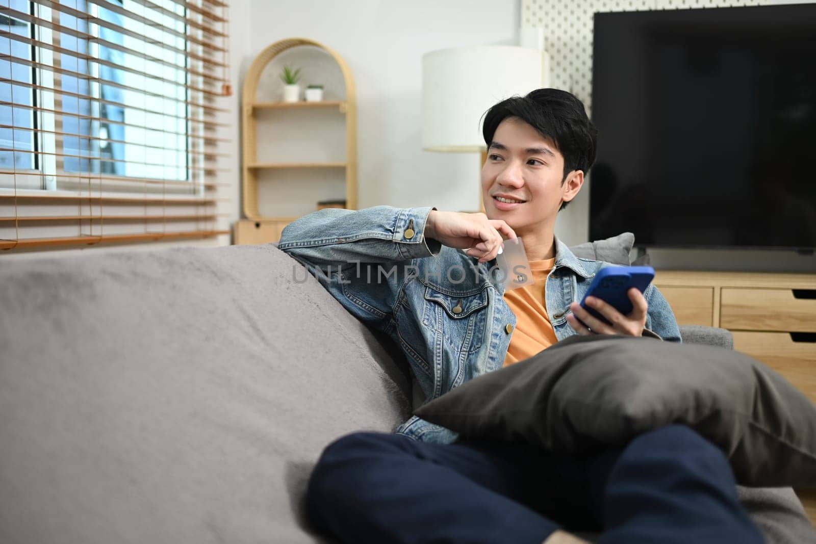 Attractive asian man relaxing on couch at home holding mobile phone and looking outside window.