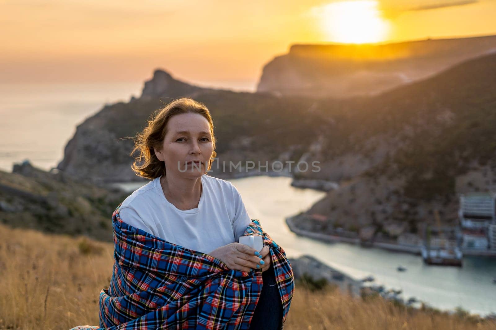 A woman is sitting on a hill with a blanket wrapped around her. She is looking out at the ocean