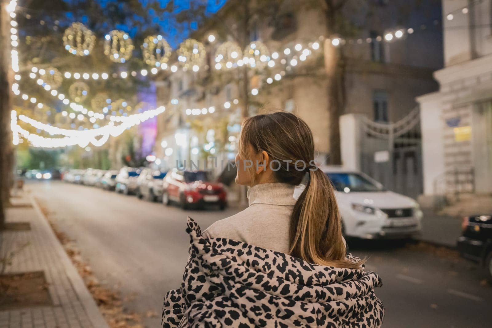 A woman wearing a leopard print coat stands on a street at night. The street is lit up with Christmas lights, creating a festive atmosphere. There are several cars parked along the street