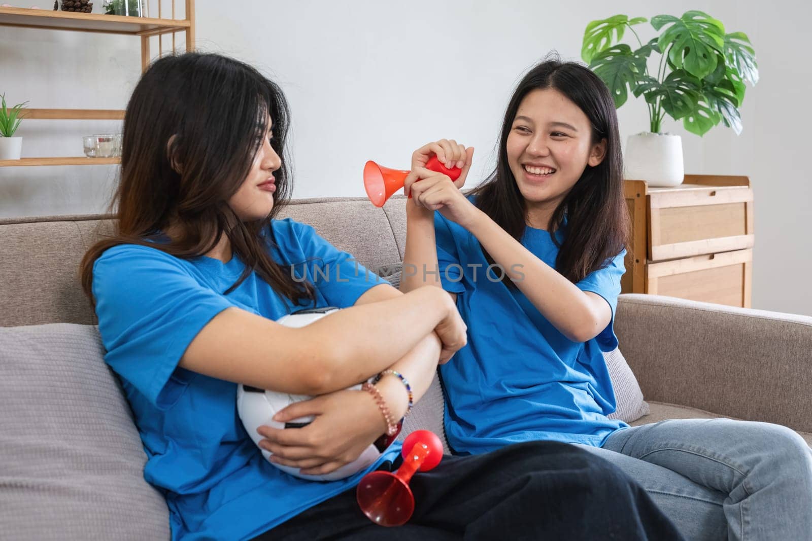 Asian women watching soccer game at home with cheering props. Concept of sports, friendship, and fun.