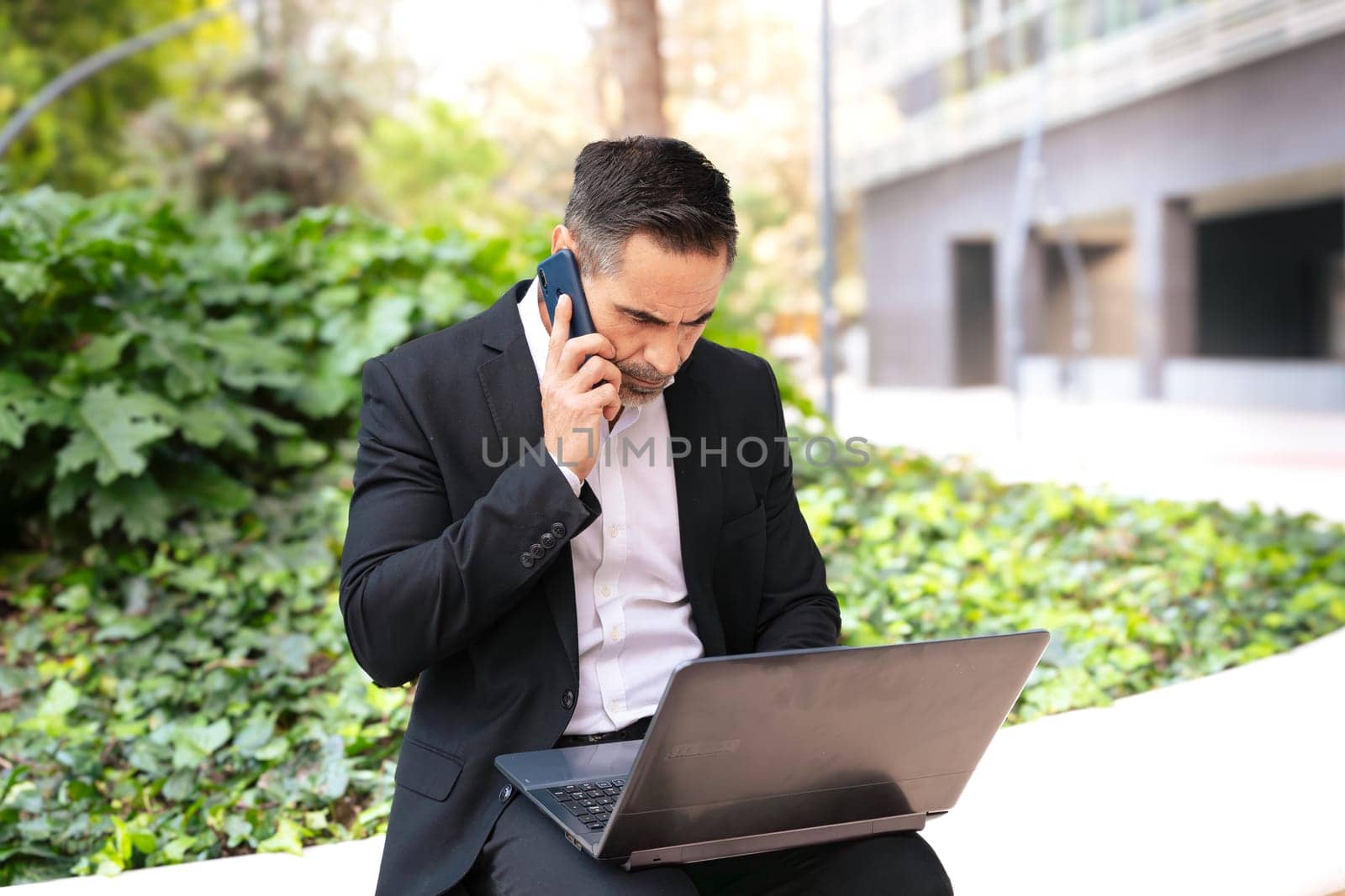 Businessman talks on a smartphone while working with laptop outdoors. by mariaphoto3