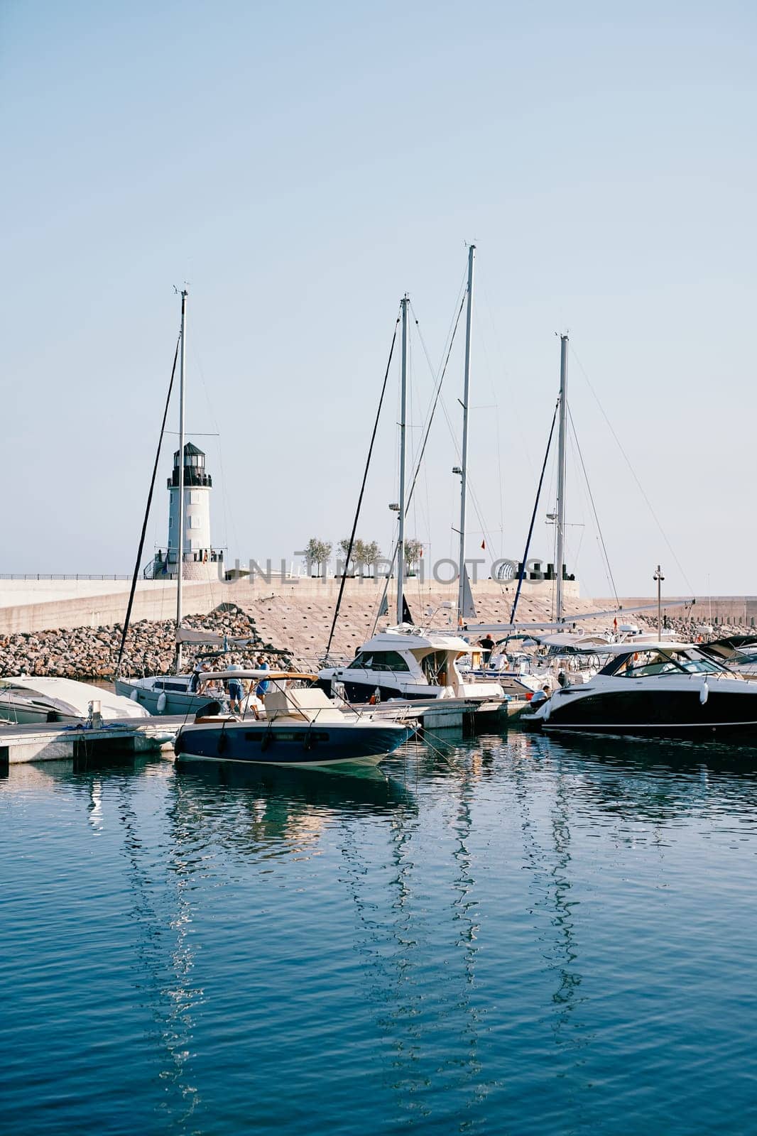 Row of motor boats moored at a breakwater with a lighthouse by Nadtochiy