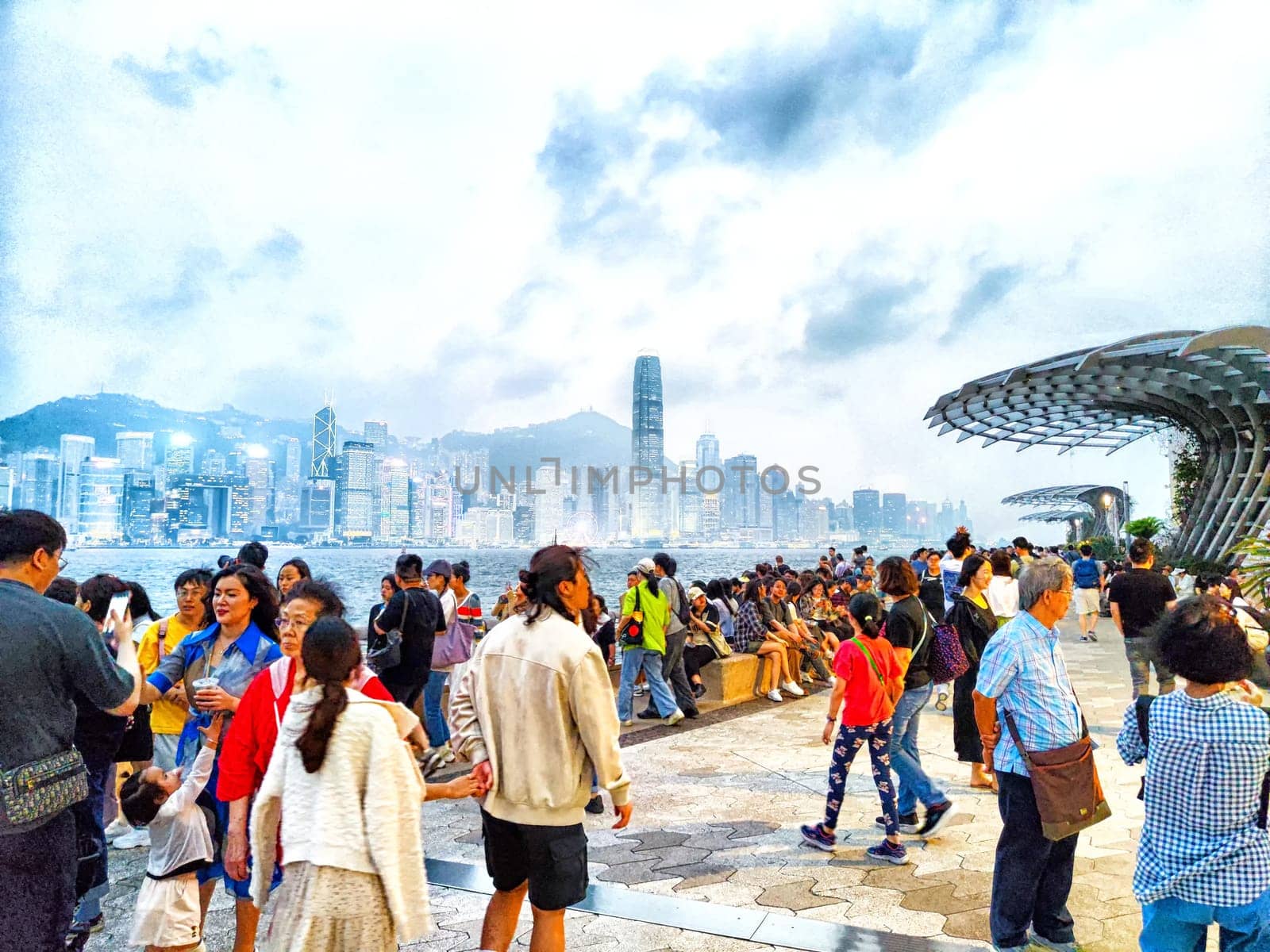 Hong Kong - April 05, 2024: People gather on the Hong Kong waterfront, with skyscrapers and Victoria Bay in the background on a cloudy afternoon