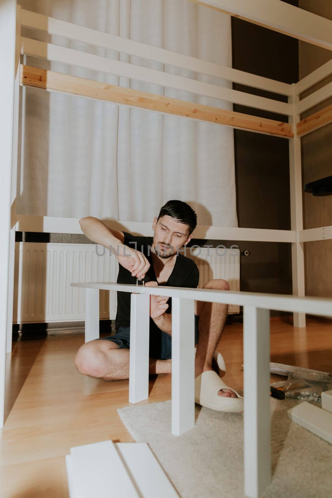 One young beautiful brunette Caucasian woman sits on the floor and tightens a screw with her hand into the hole of the white part, assembling a ladder for a bunk bed, in the evening in the room, close-up bottom view with selective focus.