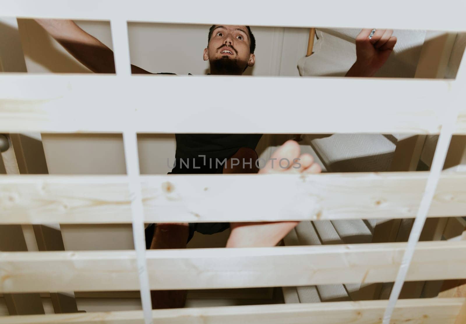 One young brunette Caucasian man installs with a bunch of frame boards on a bunk bed, in the evening in the room, close-up view from below with selective focus.