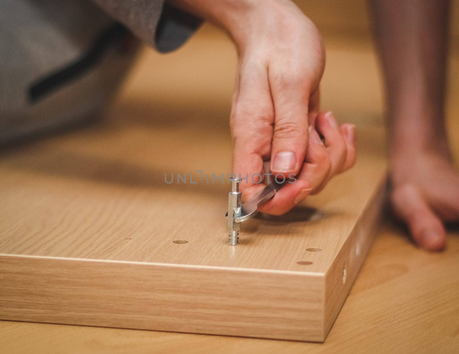 The hands of a caucasian young man with a wrench tighten a bolt on the wooden part of the bed while sitting on the floor in his room, close-up side view. The concept of assembling furniture, shopping, home. The concept of assembling furniture, shopping, at home.